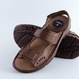 May include: Brown leather sandals with adjustable straps and a textured sole. The sandals have a round logo on the insole and are designed for comfort and support. The sole is marked with the word "SUMMER".