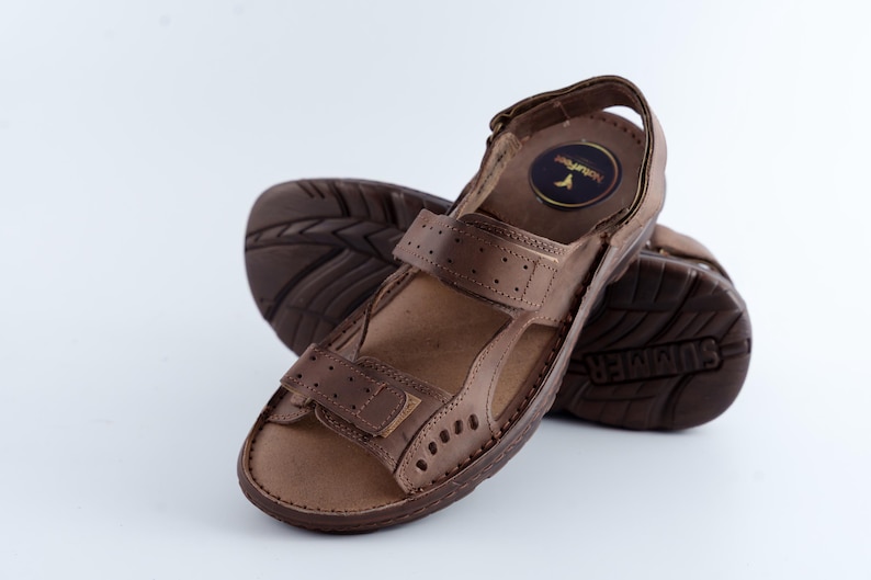 May include: Brown leather sandals with adjustable straps and a textured sole. The sandals have a round logo on the insole and are designed for comfort and support. The sole is marked with the word "SUMMER".