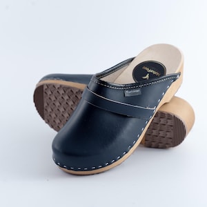Könnte beinhalten: Ein Paar marineblaue Clogs mit Holzsohle und Leder-Obermaterial. Die Clogs haben einen Riemen und weiße Nähte. Die Sohlen sind braun mit einem strukturierten Muster. Der Markenname "Naturfeet" ist sichtbar.