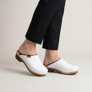 Pode incluir: Um par de tamancos de couro branco com sola de madeira clara, um acabamento castanho escuro ao longo da gola e uma pequena etiqueta preta na lateral. Os tamancos têm biqueira arredondada, design slip-on e sola castanha com rasto.