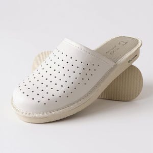 Könnte beinhalten: Weißer Leder-Slipper mit perforiertem Oberteil. Der Slipper hat eine beige Sohle und eine runde Spitze. Die Sohle ist etwa 2,5 cm dick. Der Slipper ist für den Innenbereich konzipiert.