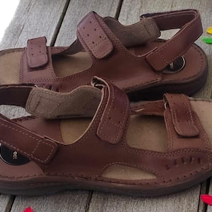 May include: A pair of brown leather sandals with adjustable straps. The sandals have a contoured footbed and are set on a wooden surface. The image also includes a potted yellow flower and some colorful petals.