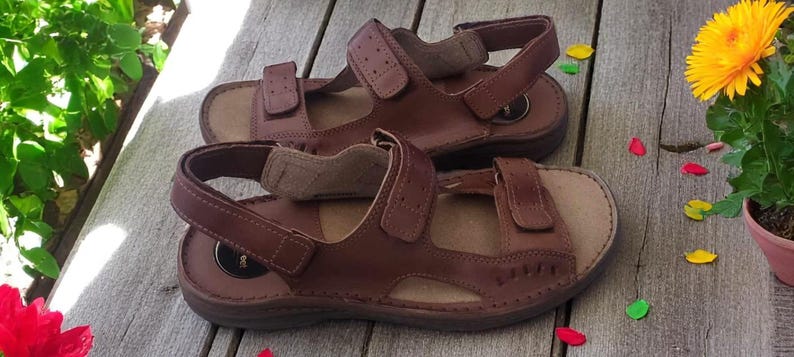 May include: A pair of brown leather sandals with adjustable straps. The sandals have a contoured footbed and are set on a wooden surface. The image also includes a potted yellow flower and some colorful petals.