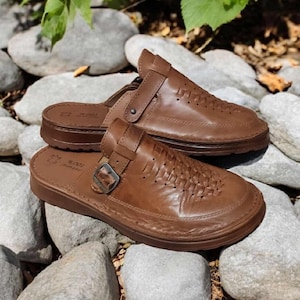 May include: A pair of brown leather sandals with a woven design and a buckle strap. The sandals have a thick sole and are sitting on a bed of rocks.