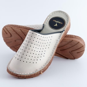 May include: Cream-colored leather slip-on clogs with a perforated upper and a brown wooden sole. The clogs have a round, closed-toe design and a stitched trim. The brand name "NaturFeet" is printed on a circular emblem.