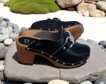 Puede incluir: Zuecos de cuero negro con tacones de madera y tachuelas plateadas. Los zuecos tienen una hebilla decorativa y se exhiben sobre una superficie de piedra. Los tacones miden aproximadamente 7,6 cm de alto.