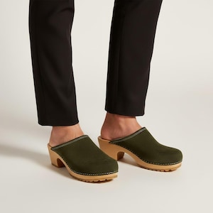May include: Olive green suede clogs featuring a rounded toe, a light-colored wooden sole with a thick heel, and visible white stitching along the upper edge where the fabric meets the wood. The clogs are worn with slim-fit black trousers.
