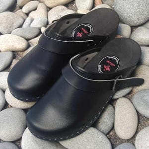 以下が含まれることがあります： 白いステッチと白いストラップが付いた、黒い革製のサボが2足。サボには、「整形外科」と「革」と書かれた赤と白のロゴがあります。サボは灰色の石のベッドの上に置かれています。
