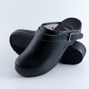 Könnte beinhalten: Schwarze Lederclogs mit Riemen und Schnalle. Die Clogs haben eine runde Spitze und eine dicke Sohle. Weiße Nähte akzentuieren die Kanten. Die Absatzhöhe beträgt ca. 5 cm.