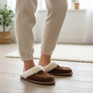Pode incluir: Chinelos de camurça castanhos com forro de pele sintética branca. Os chinelos têm um design estilo mocassim com acabamento costurado e sola de borracha. A pessoa está usando calças de moletom bege.