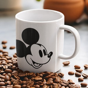 May include: A white ceramic mug with a black Mickey Mouse graphic. The mug has a curved handle and is surrounded by coffee beans. The mug is cylindrical and appears to be empty.