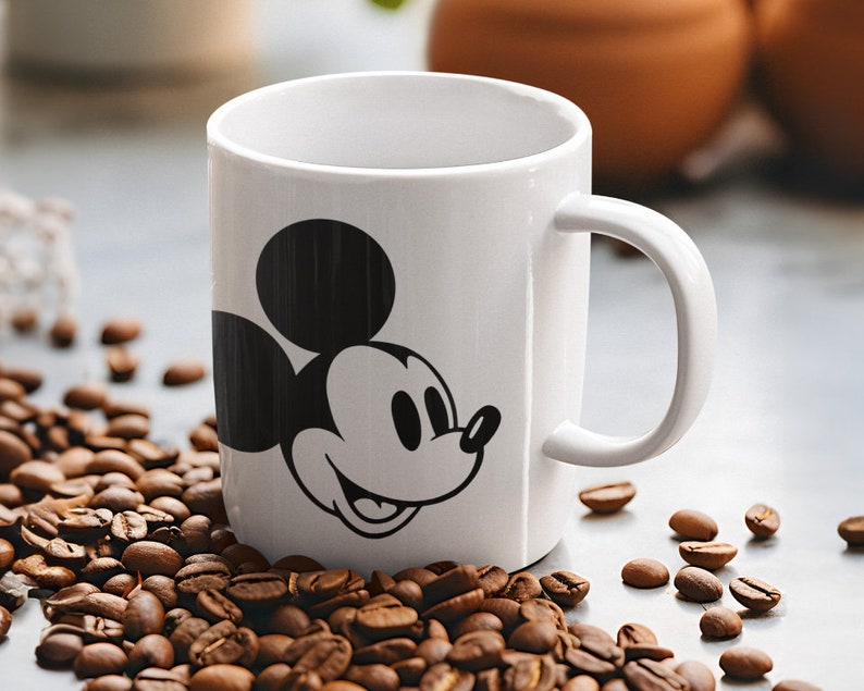 May include: A white ceramic mug with a black Mickey Mouse graphic. The mug has a curved handle and is surrounded by coffee beans. The mug is cylindrical and appears to be empty.