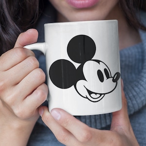 May include: White ceramic mug with a black and white Mickey Mouse illustration. The mug showcases the cartoon character's face. It has a curved handle and is held by a person. The mug is a standard size for hot beverages.