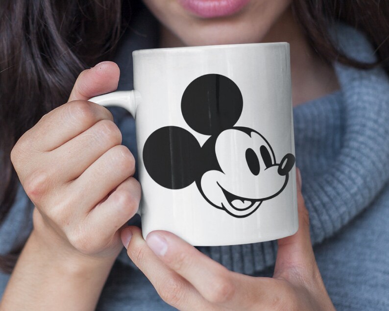 May include: White ceramic mug with a black and white Mickey Mouse illustration. The mug showcases the cartoon character's face. It has a curved handle and is held by a person. The mug is a standard size for hot beverages.