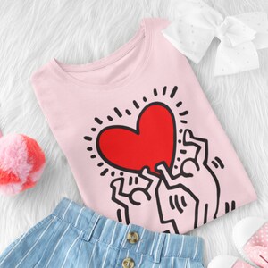 Puede incluir: Camiseta rosa con un gr&aacute;fico de tres figuras de palo sosteniendo un coraz&oacute;n rojo. El coraz&oacute;n tiene un contorno negro y est&aacute; rodeado de rayos negros.