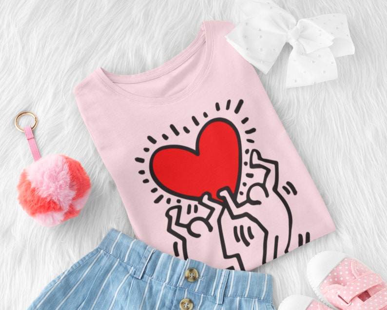 Puede incluir: Camiseta rosa con un gr&aacute;fico de tres figuras de palo sosteniendo un coraz&oacute;n rojo. El coraz&oacute;n tiene un contorno negro y est&aacute; rodeado de rayos negros.