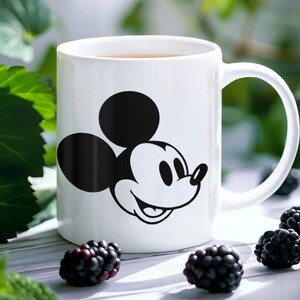 May include: White ceramic mug with a black and white Mickey Mouse design. The mug has a curved handle and contains a brown liquid. Blackberries are placed around the mug on a light surface.