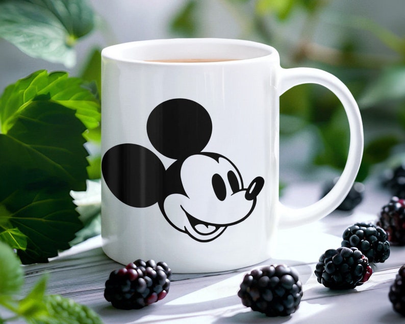 May include: White ceramic mug with a black and white Mickey Mouse design. The mug has a curved handle and contains a brown liquid. Blackberries are placed around the mug on a light surface.