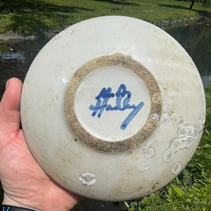 May include: A round, off-white ceramic plate viewed from the underside, featuring a circular unglazed foot ring and a blue hand-painted signature that reads Hadley. The surface shows scattered white circular spots and a smooth, glazed finish.