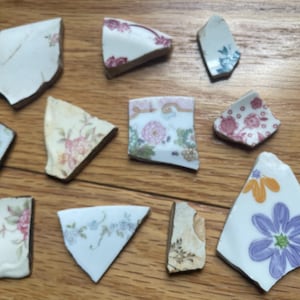 May include: A collection of broken ceramic pieces with floral patterns in various colors, including pink, blue, and yellow. The fragments are scattered on a wooden surface, showcasing the remnants of a decorative item.