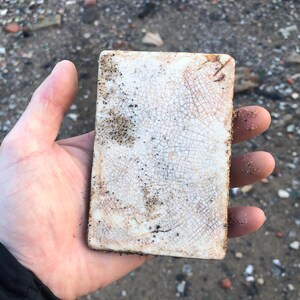 May include: A white ceramic tile with a cracked glaze and brown speckles. The tile is rectangular and has a rough texture.