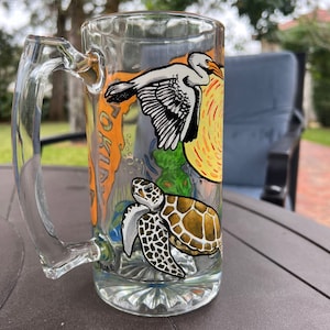 Puede incluir: Taza de cerveza de vidrio transparente con asa, con ilustraciones pintadas a mano de un pelícano, una tortuga marina y un sol amarillo. La taza tiene una base decorativa.