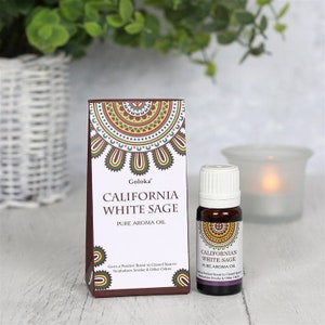 Può includere: Una scatola marrone con un design di salvia bianca e il testo "Goloka California White Sage Pure Aroma Oil" e una piccola bottiglia di vetro marrone con un tappo bianco etichettato "Californian White Sage Pure Aroma Oil".