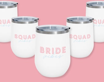 May include: Five white stainless steel wine tumblers with pink and blue lettering. The tumblers read "Bride Vibes" and "Squad Vibes".