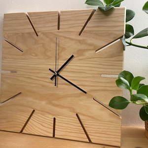 Könnte beinhalten: Eine Holzuhr mit schwarzen Zeigern und einem einzigartigen Design. Das Zifferblatt der Uhr ist aus hellem Holz mit einer Maserung und hat eine quadratische Form mit Ausschnitten an den Rändern.