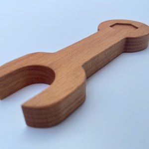 Può includere: Una chiave inglese giocattolo in legno di colore marrone rossiccio caldo. Il giocattolo presenta la classica forma di una chiave inglese con un manico arrotondato e ganasce aperte. La venatura del legno è visibile, aggiungendo consistenza. Il giocattolo è per bambini.