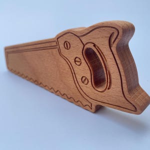 Könnte beinhalten: Eine Holzhandsäge mit detailliertem Griff und Sägeblatt. Die Säge ist aus hellbraunem Holz gefertigt und weist ein geschnitztes Design auf. Das Sägeblatt hat eine Reihe von Zähnen entlang der Unterkante.