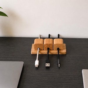 Op de afbeelding: Een houten kabelorganizer met drie vierkante blokken en een rechthoekige basis. Elk blok heeft een kabel: een witte USB-C, een zwarte USB en een zwarte audio jack. De organizer is gemaakt van lichtgekleurd hout.