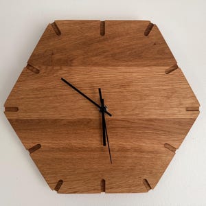 May include: A hexagonal wooden wall clock. The clock face is made of light brown wood with visible grain. Black clock hands show the time. The clock has small notches around the edges. The clock's dimensions are approximately 30 cm.