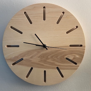 Könnte beinhalten: Eine runde Holzuhr mit hellem Holzton. Das Zifferblatt hat gleichmäßig verteilte, längliche Aussparungen am Rand, die als Stundenmarkierungen dienen. Schwarze Zeiger zeigen die Uhrzeit an. Die Uhr ist an einer hellen Wand befestigt.