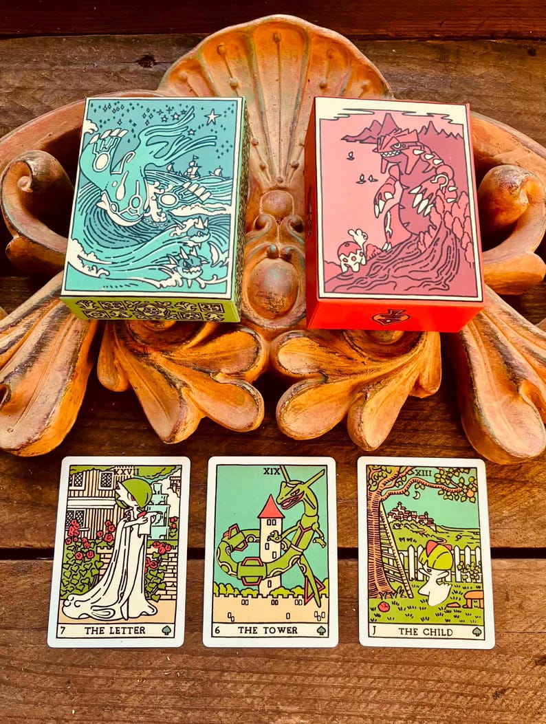 K&ouml;nnte beinhalten: Zwei rechteckige Kartenboxen mit t&uuml;rkisfarbenen und rosafarbenen Kreaturendarstellungen, begleitet von drei Tarot-Karten mit den Aufschriften The Letter, The Tower und The Child, die jeweils eine Figur in einer Szenerie zeigen.