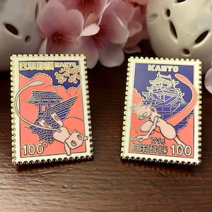May include: Two enamel pins with a gold border, featuring a pink and blue design with a cartoon character and Japanese text. One pin reads "KANTO" and "100". The other pin has the same design and reads "100".