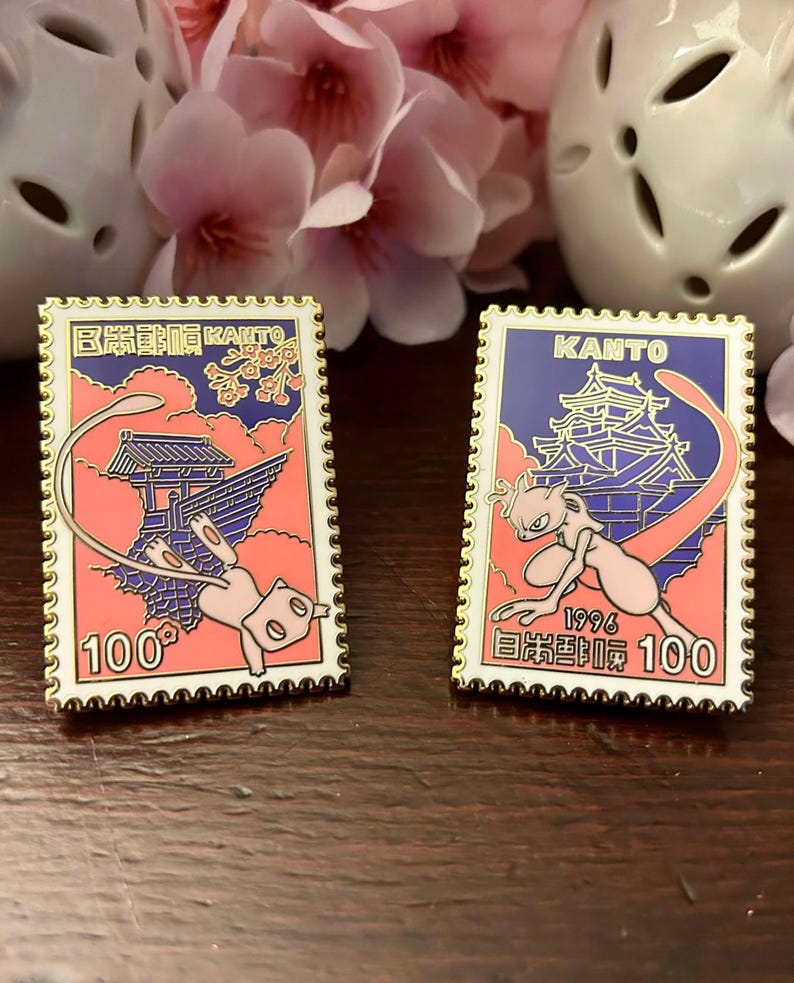 May include: Two enamel pins with a gold border, featuring a pink and blue design with a cartoon character and Japanese text. One pin reads "KANTO" and "100". The other pin has the same design and reads "100".