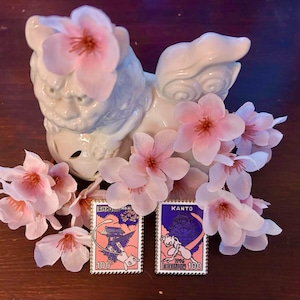 May include: Two collectible pins featuring cartoon characters, set against a backdrop of pink cherry blossoms and a white ceramic guardian statue. The pins have a postage stamp design with the text "100" and Japanese text.