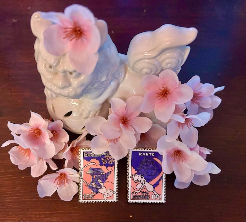 May include: Two collectible pins featuring cartoon characters, set against a backdrop of pink cherry blossoms and a white ceramic guardian statue. The pins have a postage stamp design with the text "100" and Japanese text.