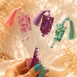 Peut inclure: Trois marque-pages en résine transparente avec des paillettes colorées et des pompons. Les marque-pages présentent des paillettes roses, violettes et turquoise, chacun avec un pompon assorti. Les marque-pages sont tenus dans une main.