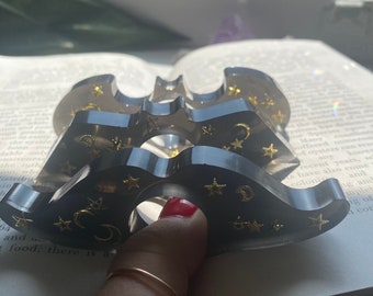 Könnte beinhalten: Ein dunkelgraues, fledermausförmiges Objekt aus Kunstharz mit goldenen Sternen und Mondakzenten. Das Objekt wird über einem offenen Buch gehalten. Das Harz ist durchscheinend. Das Design dient wahrscheinlich dekorativen oder spirituellen Zwecken.