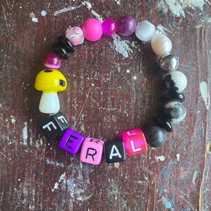 May include: A colorful beaded bracelet with a mushroom charm and letter beads spelling out "FERAL". The bracelet features black, white, pink, purple, and yellow beads.