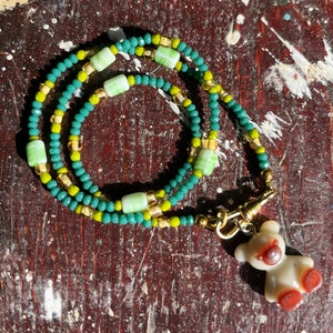 Può includere: Una collana di perline verdi e dorate con una perlina rettangolare verde e bianca e un ciondolo a forma di orso in ceramica bianca con dettagli marroni.