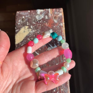 Puede incluir: Una pulsera de cuentas coloridas con un dije de mariposa rosa. La pulsera presenta cuentas en tonos rosa, blanco, turquesa y verde. El dije de mariposa es rosa translúcido con detalles brillantes.