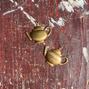 May include: Two gold-coloured teapot charms on a brown wooden surface. The charms are detailed with a spout, handle, and lid.