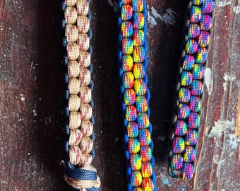 Puede incluir: Tres llaveros de paracord coloridos con llaveros de oro y plata. Los llaveros están hechos con un patrón tejido y tienen diferentes combinaciones de colores. Los llaveros están sobre una superficie de madera.