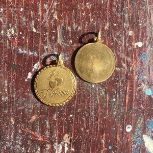 May include: Gold-toned pendant with a heart design and the inscription "Je t'aime" on one side and a plain, smooth surface on the other side.