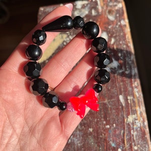 Puede incluir: Pulsera de cuentas negras con un dije de lazo rojo. Las cuentas son facetadas y tienen un acabado brillante.
