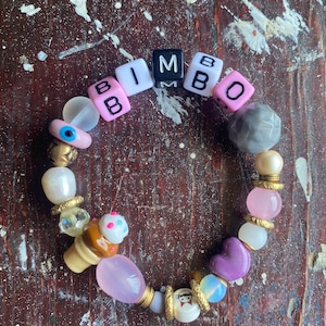 May include: A colorful beaded bracelet with the word "BIMBO" spelled out in pink, white, and black letter beads. The bracelet also features a variety of other beads, including an evil eye bead, a heart bead, and a bead shaped like an ice cream cone.