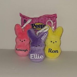May include: Three plush Easter bunny ornaments in pink, purple, and yellow, each with a name embroidered on the front. A package of Peeps marshmallows is in the background. The purple bunny has "2022" embroidered on it.
